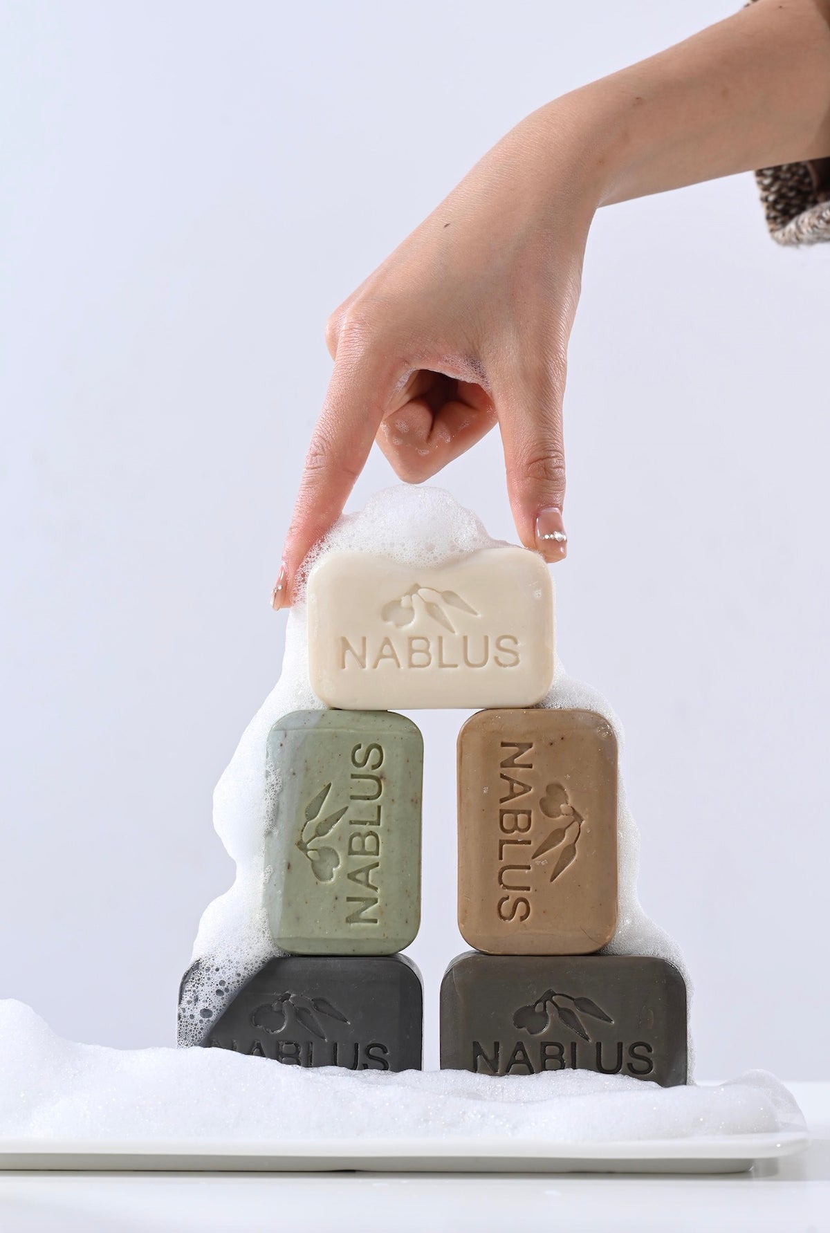 Hand reaching for a bar of Nablus soap among other bars on a white surface.
